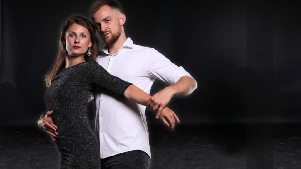 Taneční pár elegantně pózuje v těsném objetí, žena ve třpytivých černých šatech a muž v bílé košili a černých kalhotách, proti tmavému pozadí. Učitelé taneční lekce pro začátečníky.