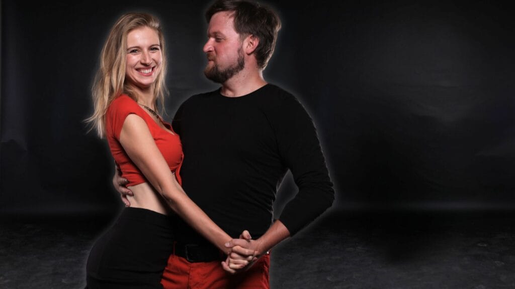 Sebevědomý taneční pár pózující proti tmavému pozadí; žena v červeném crop topu a černé sukni, muž v černém tričku s červenými kalhotami, v těsném tanečním objetí.