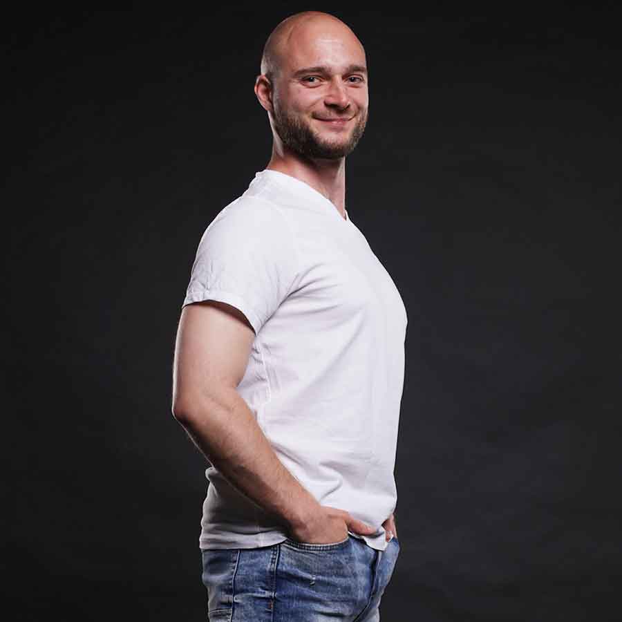 Mužský učitel salsy a bachaty v bílém tričku, stojící sebevědomě s klidným úsměvem a rukama na bocích.