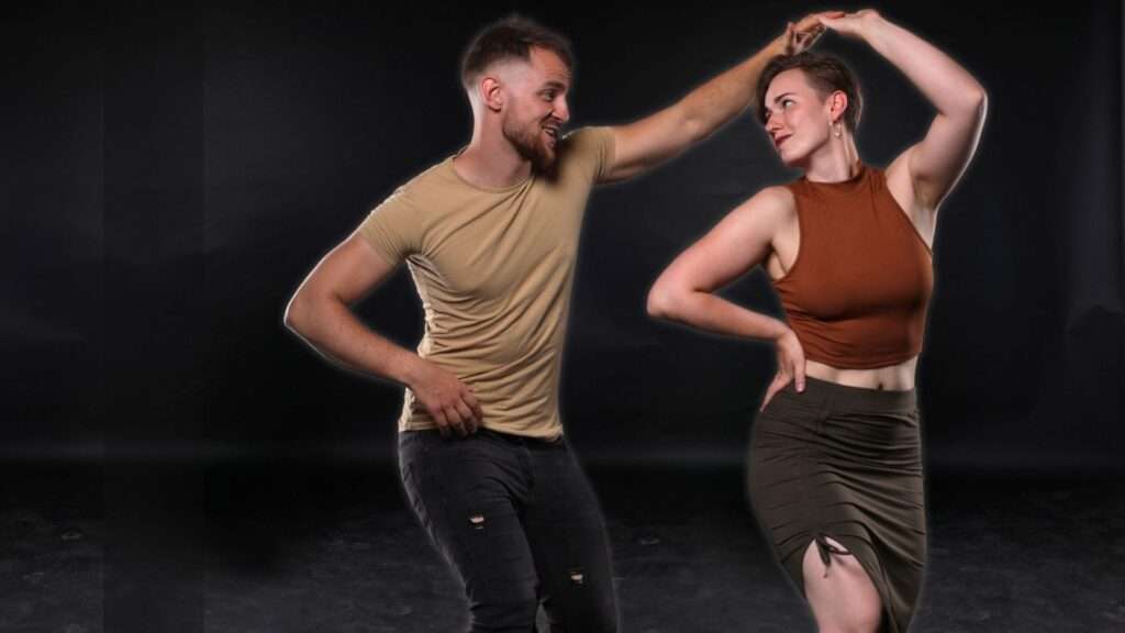 Radostný taneční pár předvádějící hravý salsa krok. Mužský tanečník v béžovém tričku a tmavých džínách vede svou partnerku s jistotou, zatímco tanečnice v hnědém topu a olivové sukni následuje s grácií a stylem. Jejich výrazy odrážejí radost a energii tance na elegantním tmavém pozadí.